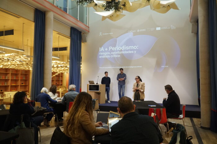 Personas asistentes a un taller titulado "IA + Periodismo: riesgos, oportunidades y análisis crítico", donde tres ponentes presentan el tema en un espacio moderno con una gran pantalla proyectando la información.